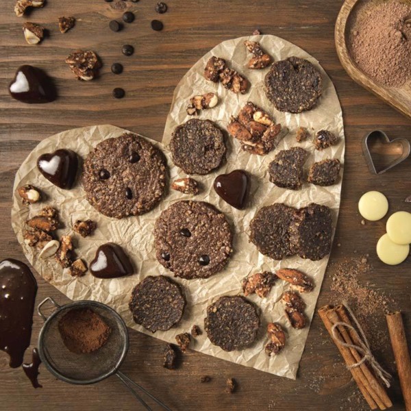 Image shows a heart shaped parchment paper with cookies, buts, granola and chocolates on top. Parchment paper is on top of a wooden background and has spices, chocolate and cinnamon surrounding the heart.
