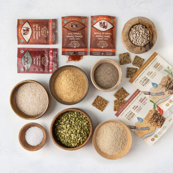 Image shows two bags of KETO U Bake cracker mixes with ready made crackers spilling out of them and ingredients in various sized wooden bowls (chia seeds, pumpkin seeds, sunflower seeds, flax) as well as two red bags of Gathering Place Trading spices and herbs