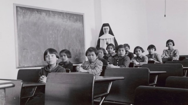Inside the classroom of an Indian Residential School, school children are sitting at desks while a nun looks on in the background