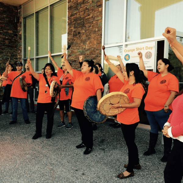 Group of Indigenous people gathered together wearing orange shirts and holding traditional drums.