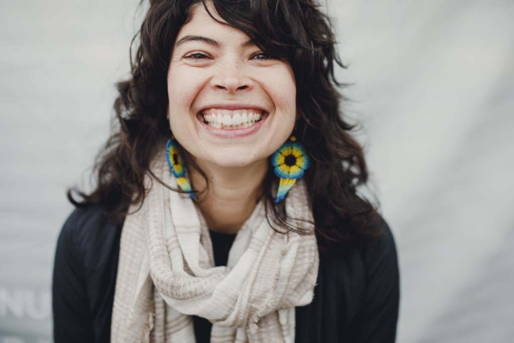 Practice With Clara, image of Clara smiling with long dark hair and colourful blue and yellow earrings. She is wearing a dark long sleeved shirt and a light scarf around her neck.